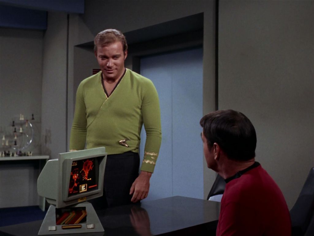 A scene from the Star Trek episode The Trouble with Tribbles. Kirk, standing, is speaking with a seated Scotty. On the screen in front of Scotty is an engineering schematic. 